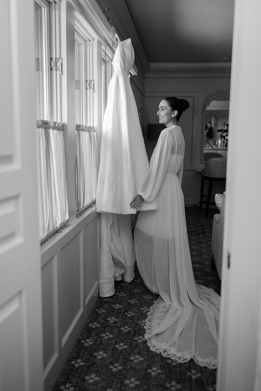 Black and white film photo of the bride smiling warmly at her hanging wedding gown by a window.